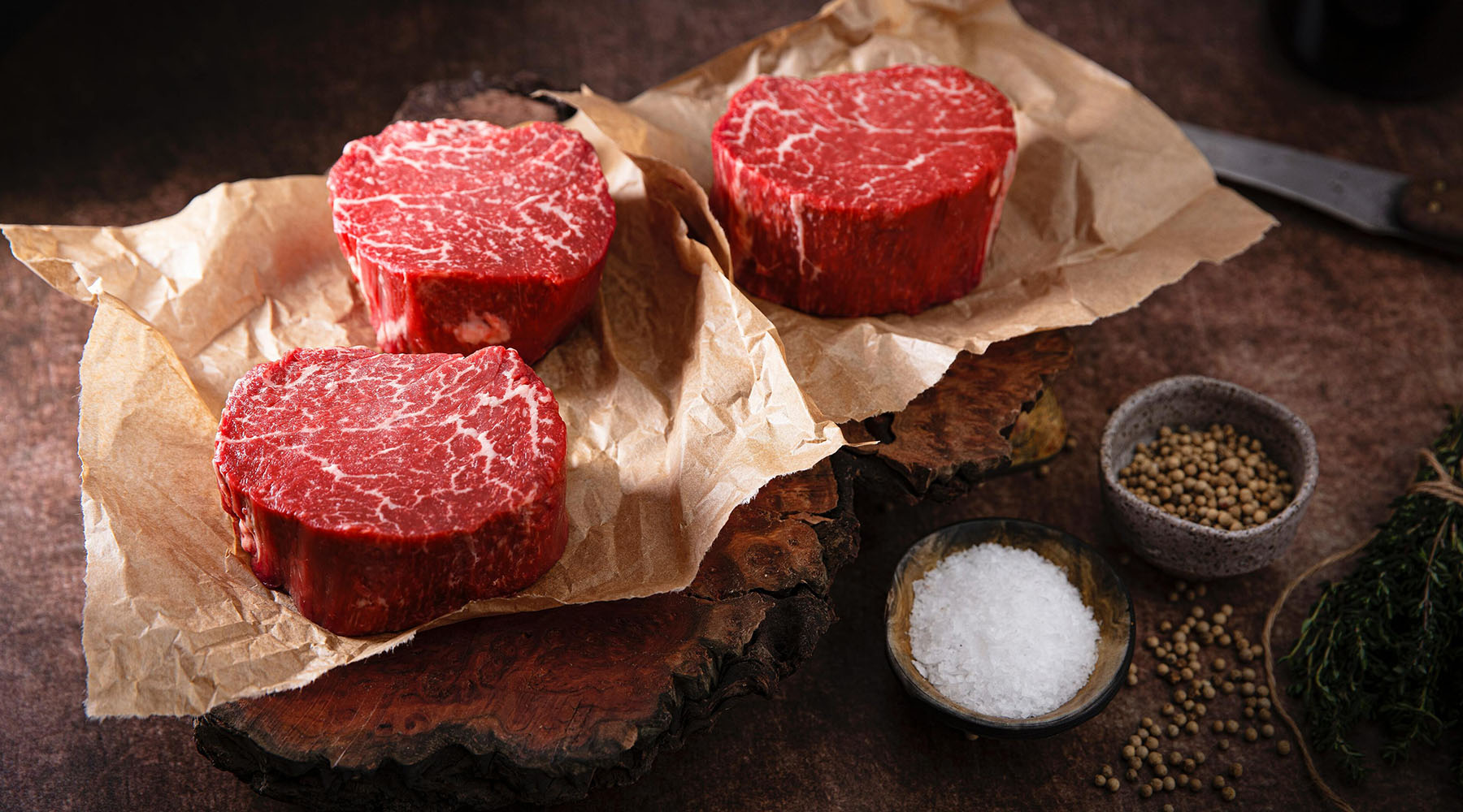 Three raw grass-fed beef tenderloin steaks with marbling, arranged on brown butcher paper over a rustic wooden board, with small bowls of salt, peppercorns, and herbs nearby on a dark tabletop.