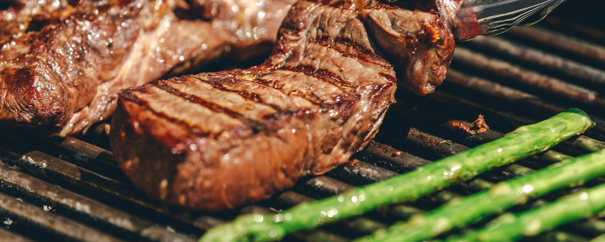 Grilled steak with visible grill marks cooking on a grill grate, with green asparagus in the foreground and a basting brush at the top right.