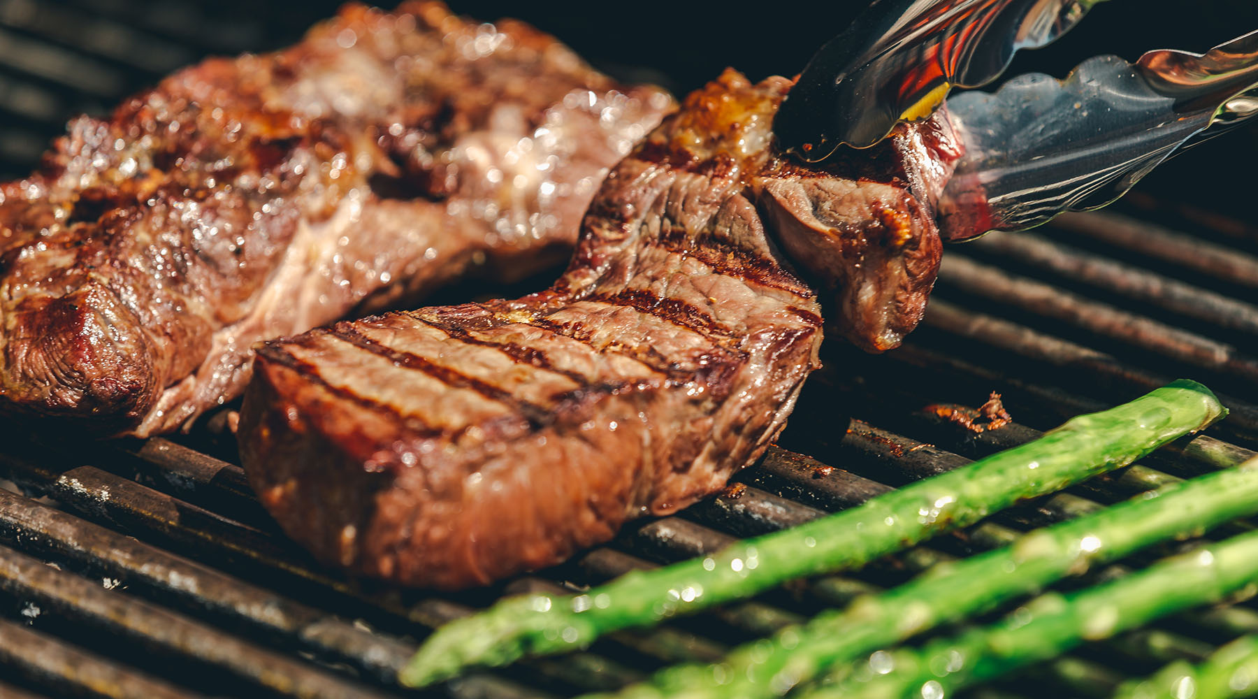 Grass-fed rilled steak on a barbecue grill with visible grill marks, alongside green asparagus and a pair of tongs holding a piece of meat.
